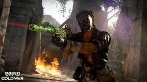 A soldier with a tribal mask on aims a LC10 rifle with a laser on it while moving through ruins with a fire behind him. In the corner is the Call of Duty Black Ops Cold War logo.