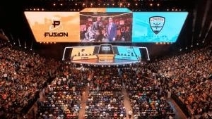 A stadium is filled with people there to spectate an Overwatch League match. Giant monitors are hoisted from the ceiling and display the Philadelphia Fusion and London Spitfire logos with the middle screen showing an interview with a player.