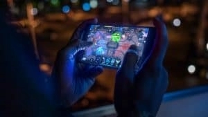 A person plays a mobile game on their smartphone on a balcony in the city at night.