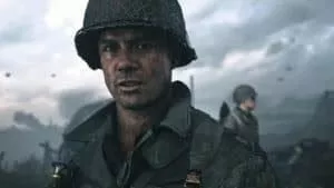 A close up of a soldier's face, under his helmet as he stands amid a battlefield looking at the camera